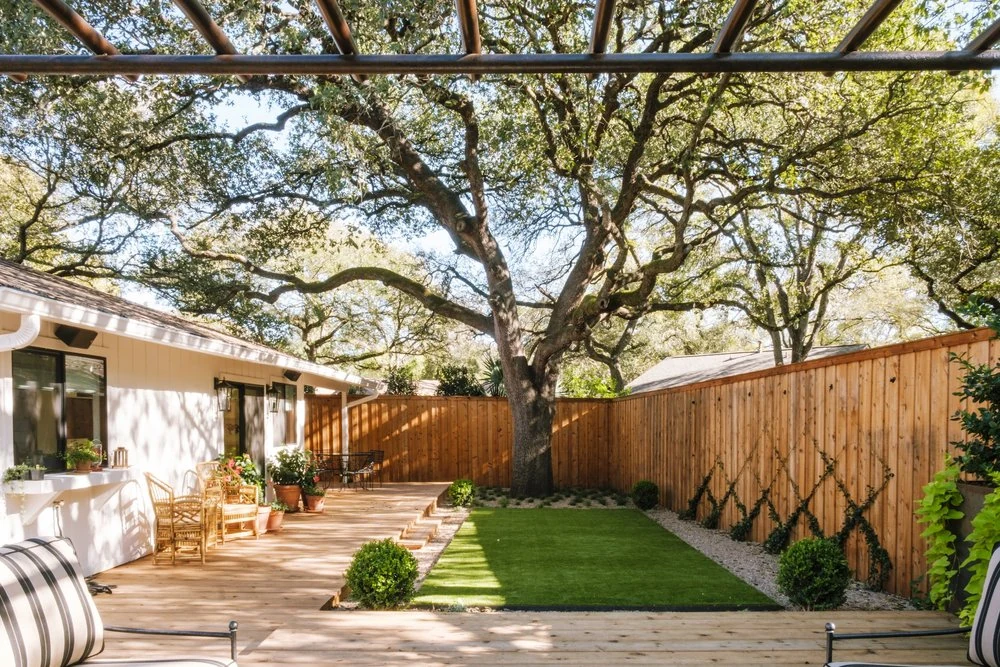 A wood patio and lawn enclosed by a tall wood fence beneath a mature shade tree