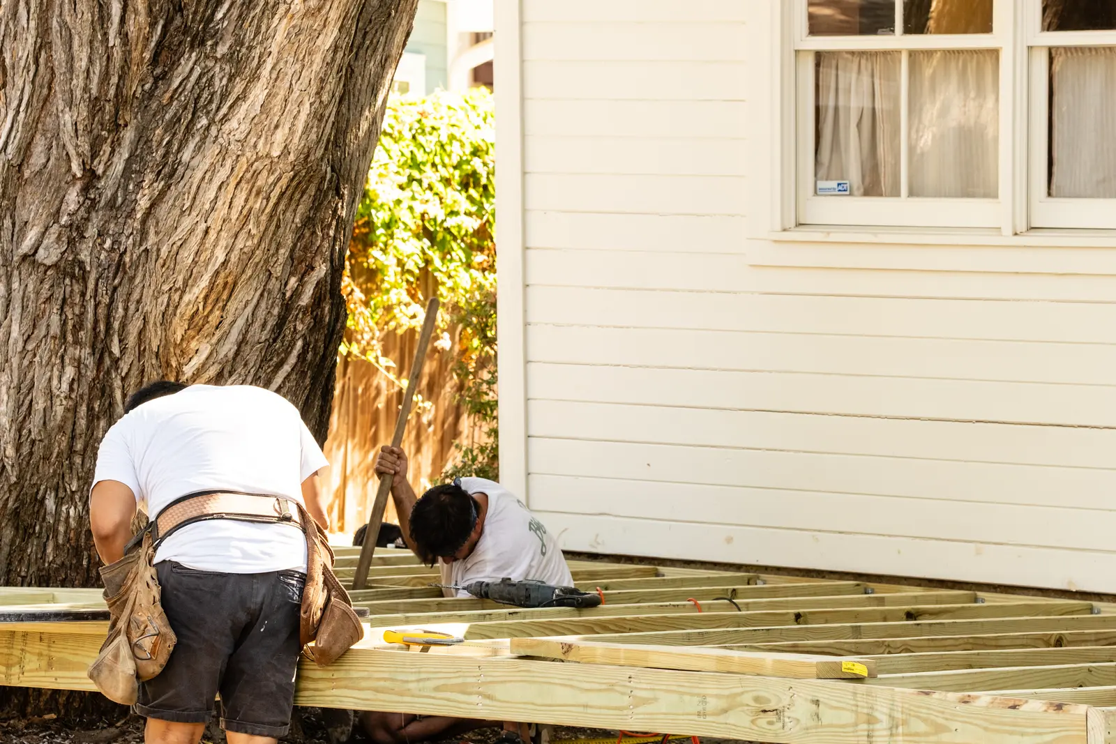 Two crew members fitting deck framing beside a mature tree and a white house