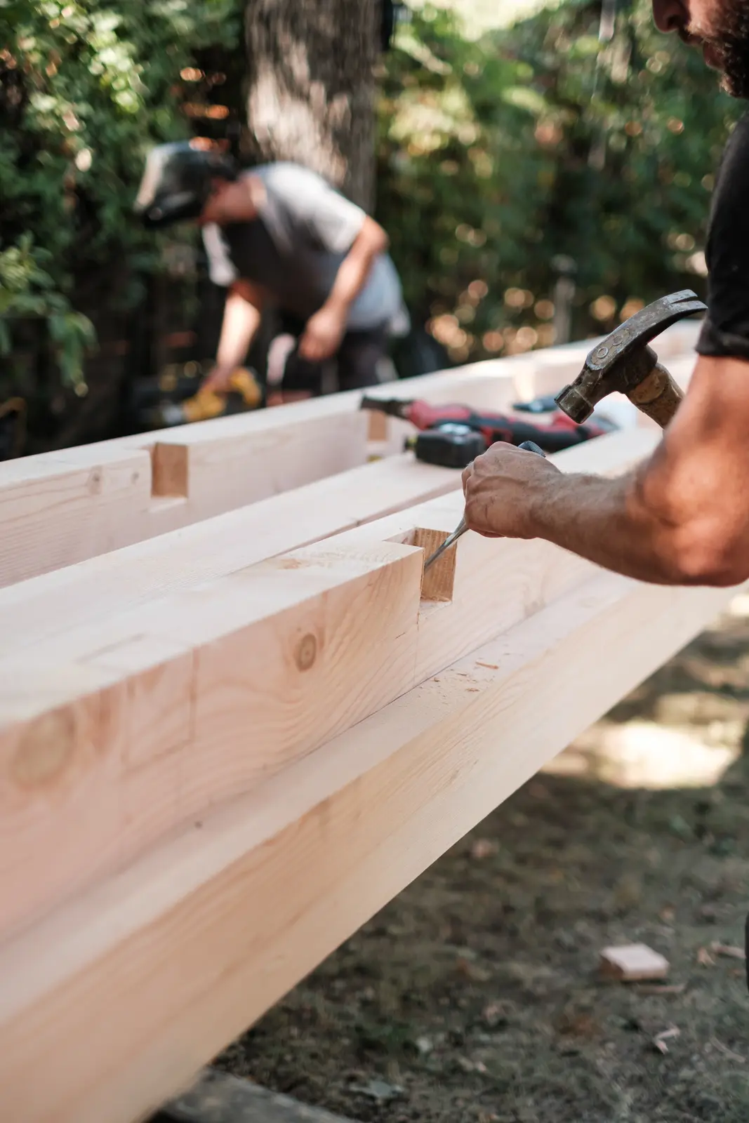 A carpenter chiseling a timber joint with another crew member working in the background