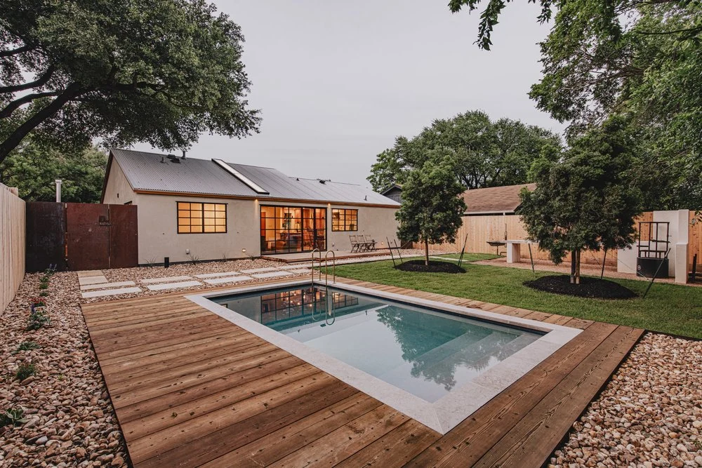 A wood deck wrapping around a rectangular pool in a landscaped backyard